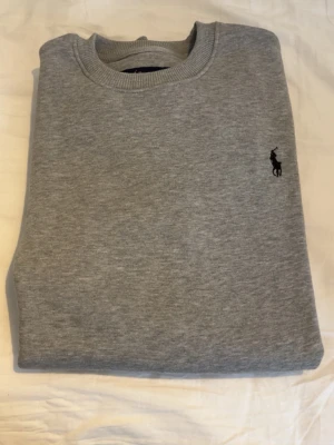 Grå sweatshirt från Polo Ralph Lauren - Klassisk grå sweatshirt från Polo Ralph Lauren med rund halsringning och broderad logga på bröstet. Tröjan är i mjukt material och har ribbade muddar vid hals, ärmslut och nederkant. Perfekt för en chill och stilren look. Kontakta för intresse/frågor!