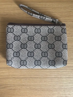 Beige mönstrad plånbok med handledsrem - Snygg beige/brun plånbok med mörkgrått mönster. Trendig och fin. Plånboken har dragkedja upptill. Perfekt för att bära det viktigaste, kort, smink el annat och är bra att ha i en större väskan, för orningens skull.  (Handkrämen ingår ejj 