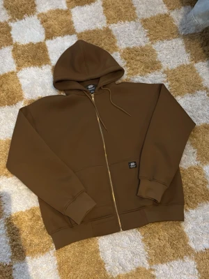 Sweet sktbs zip hoodie - Säljer en brun hoodie med dragkedja och huva. Hoodien har ribbade muddar, fickor framtill och en liten svart patch på ena sidan. Perfekt för en chill och avslappnad stil. Materialet känns mjukt och skönt mot huden.