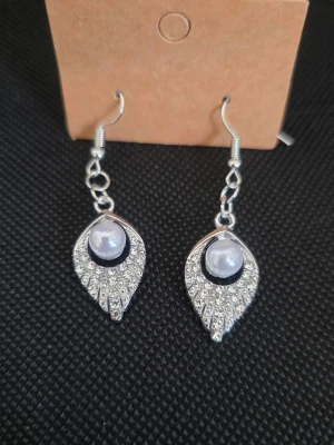 Silvriga örhängen med pärldetalj - Snygga hängande örhängen i silverfärg med glittrande stenar och en vit pärla i mitten. Formen är droppformad och örhängena har en elegant och modern design. Perfekta för att lyfta din stil med lite extra bling.