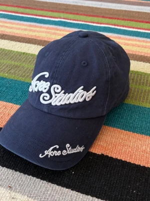 Mörkblå keps från Acne Studios - Snygg mörkblå keps från Acne Studios med broderad logga i vitt framtill och på skärmen. Klassisk böjd skärm och justerbar baksida. Tillverkad i bomull för en bekväm och avslappnad look.