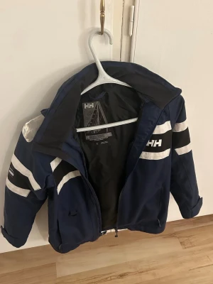 Marinblå vindjacka från Helly Hansen - Snygg marinblå vindjacka från Helly Hansen med vita ränder på ärmarna och logga på bröstet. Jackan har dragkedja framtill, hög krage och långa ärmar. Perfekt för blåsiga dagar och har en sportig look.
