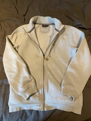 Ljusgrå zip hoodie från Sail Racing - Ljusgrå hoodie från Sail Racing med dragkedja framtill och ståkrage med svartvit randig detalj. Tillverkad i mjukt material och har diskret Sail Racing-logga på bröstet. Perfekt för en avslappnad och sportig stil.