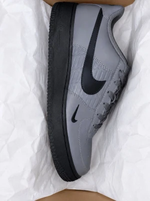 Nike Air Force 1 grå/svarta sneakers - Nike Air Force 1 sneakers i grått med svart swoosh och svart sula. Klassisk låg modell med ovandel i läder och snygga detaljer längs sidan. Perfekta för dig som vill ha en clean och stilren look med streetkänsla.
