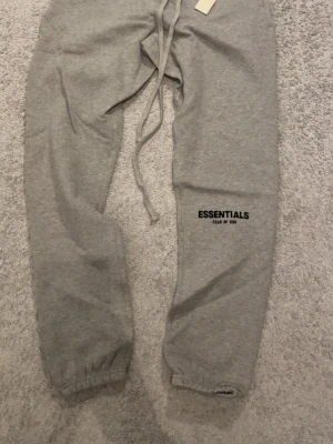 Grå Essentials Fear of God mjukisbyxor - Säljer ett par grå mjukisbyxor från Essentials Fear of God. Byxorna har resår i midjan och benslut, samt långa dragsnören och logga tryckt på benet. Tillverkade i mjukt sweatshirtmaterial som är perfekt för chill dagar.