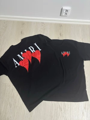  2 ST Svart AMIRI t-shirt med röda hjärtan -  2 ST Svart t-shirt från AMIRI med tryck av tre röda hjärtan och vit logga både fram och bak. Klassisk rund hals och korta ärmar. Perfekt statement-plagg för dig som gillar streetwear och snygga detaljer.