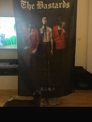 Palaye Royale The Bastards World Tour 2020 tygposter - Stor svart tygposter med motiv från Palaye Royale The Bastards World Tour 2020. Trycket visar tre personer, två i röda kavajer och en bar överkropp, samt texten 'The Bastards' högst upp. Perfekt för att dekorera väggen med bandtema.