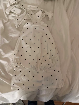 Vit prickig axelbandslös topp - Säljer en vit topp med svarta prickar och knytband i nacken. Toppen är axelbandslös och har en croppad passform med resår i midjan. Perfekt för dig som gillar en lekfull och trendig stil. Materialet känns som bomull eller polyester.