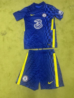 Chelsea FC matchställ blå/gul Nike - Säljer ett Chelsea FC matchställ med tröja och shorts i blått med gula detaljer. Tröjan har klubbmärke, sponsorlogga och Nike-logga, samt mönster i zigzag och rutor. Shortsen har elastisk midja, klubbmärke och gula ränder på sidorna. Perfekt för fotbollsträning!