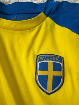 Sverige matchtröja i gult och blått - Gul och blå matchtröja från SOC med Sveriges landslagsemblem på bröstet. Tröjan har korta ärmar och är tillverkad i ett lätt och funktionellt material, perfekt för fotboll eller träning. Snygg design med blå detaljer längs sidor och axlar. Strl 146-152 men passar mig perfekt som bär Xs-S
