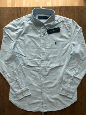 Ljusblå skjorta från Ralph Lauren - Klassisk ljusblå skjorta från Ralph Lauren med button-down krage och broderad logga på bröstet. Skjortan har långa ärmar, vita knappar och är tillverkad i mjuk bomull. Perfekt för en clean och stilren look.
