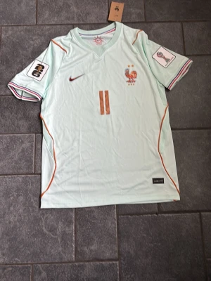 Ljusblå Frankrike fotbollströja - Säljer en ljusblå fotbollströja med bronsfärgade detaljer och nummer 11, samt namnet Olise på ryggen. Tröjan har Frankrikes landslagsemblem och Nike-logga på bröstet, samt broderade märken på ärmarna. Tillverkad i ett lätt och ventilerande material.