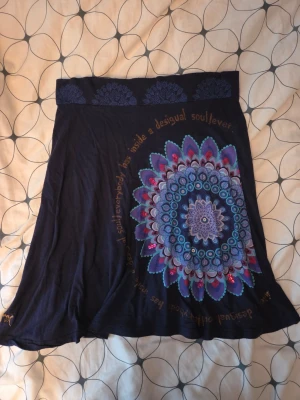 Vintage Mörkblå kjol med mandalamönster Desigual - Unik mörkblå kjol från Desigual i mjukt material. Kjolen har ett stort färgstarkt mandalamönster i blått, lila och rosa samt textdetaljer runt om. Perfekt för dig som gillar boho-vibbar och vill sticka ut lite extra.