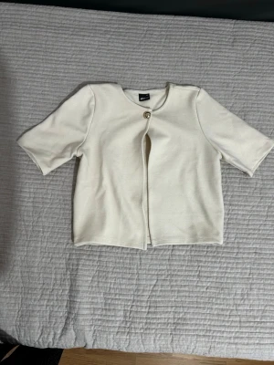 Kortärmad vit kofta med knapp - Stilren vit kofta med korta ärmar och en dekorativ knapp framtill. Koftan har rak passform och är tillverkad i ett mjukt, stickat material. Perfekt att slänga över en topp för en clean look.