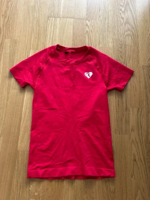 Röd t-shirt med hjärtdetalj - Snygg röd träning t-shirt med korta ärmar och en vit hjärtdetalj på bröstet. T-shirten har en klassisk rund halsringning och är tillverkad i mjukt bomullstyg. Perfekt för dig som gillar färg och vill sticka ut lite extra.