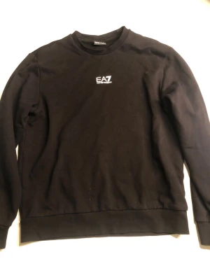 Svart sweatshirt från EA7 Emporio Armani - Snygg svart sweatshirt från EA7 Emporio Armani med vit logga på bröstet. Tröjan har rund halsringning, långa ärmar och ribbade muddar vid ärmslut och nederkant. Perfekt för en clean och stilren look.