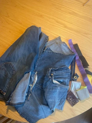 Jeansmaterial och tygbitar i blått - Ett paket med återbrukat jeansmaterial i olika blå nyanser, inklusive fickor, dragkedjor och tygbitar. Perfekt för DIY-projekt, lagning eller kreativa syprojekt. Innehåller även några färgglada tygremsor och detaljer.