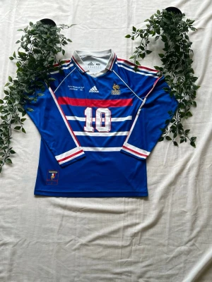 Blå Frankrike fotbollströja Zidane - Klassisk blå långärmad Frankrike fotbollströja från Adidas med nummer 10 och Zidane på ryggen. Tröjan har vita och röda detaljer, broderad tupp-logga och FIFA World Cup 1998-tryck på bröstet. Perfekt för dig som älskar retro och fotboll.