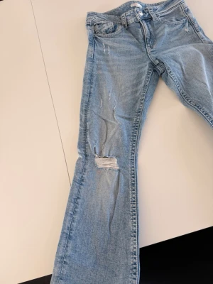Ljusblå slitna bootcut jeans - Säljer ett par ljusblå jeans med bootcut-modell och slitningar på benen, inklusive en större reva på vänster lår. Jeansen har klassiska fickor fram och bak med lock och knapp. Perfekta för en avslappnad och trendig look. Säljer då dom inte kommer till användning (säljs inte längre) 