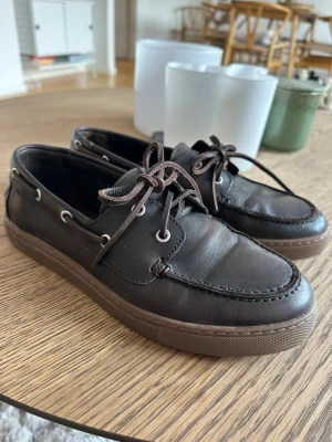 Bruna seglarskor från K.Cobler - Mörkbruna seglarskor i läder från K.Cobler med klassisk snörning och kontrastsömmar. Skorna har en robust gummisula och metallöglor för snörena. Perfekta för en avslappnad och stilren look. Använda ett fåtal gånger pga något för små för mig. Nypris 1400kr
