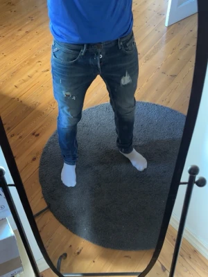 Blå slitna jeans från Replay - Säljer ett par blå jeans från Replay med slitningar och hål på låren. Jeansen har fem fickor och klassisk knappgylf. Snygg tvätt med ljusare partier och detaljerade sömmar. Perfekta för en avslappnad och trendig look.