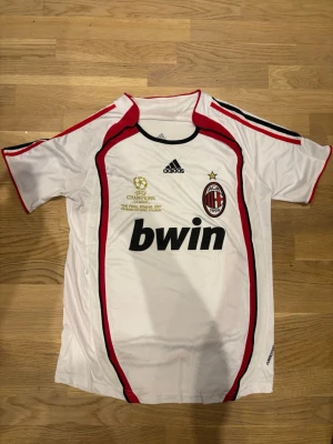 AC Milan Champions League tröja 2007 - Vit AC Milan tröja från Adidas med röda och svarta detaljer, Champions League-finalen 2007 broderat i guld, klubbmärke och sponsortryck. Korta ärmar och sportig passform, perfekt för fotbollsfans.