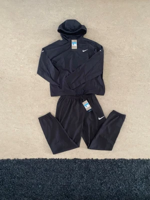 Nike Miller set - Snyggt svart träningsset från Nike med matchande hoodie och byxor. Setet har Nike-logga på både tröja och byxa, huva på tröjan och resår i midjan på byxorna. Materialet är lätt och andas bra. Storlek M. Perfekt för gym, träning eller vardagsoutfit.