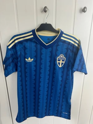 Blå Sverige bortatröja 2026 - Säljer en blå fotbollströja med Sveriges landslagsemblem på bröstet och guldfärgade ränder på axlarna. Tröjan har ett retroinspirerat mönster, V-ringning och korta ärmar. Materialet är lätt och andas, perfekt för träning eller match eller i vardagen.