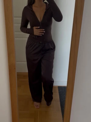 Brun träningströja - Säljer ett brunt loungewear-set med långärmad topp och dragkedja framtill samt matchande byxor med resår i midjan. Setet har en mjuk och bekväm passform, perfekt för chill dagar hemma. Materialet ser ut att vara stretchigt och följsamt.