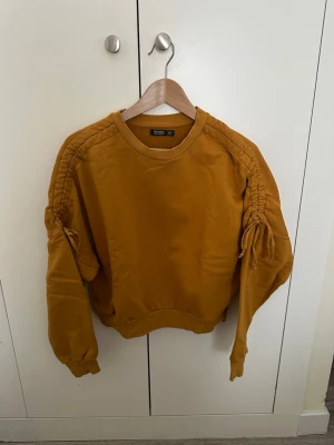Gul sweatshirt med snörning från Bershka - Cool gul sweatshirt från Bershka med snörningsdetaljer längs ärmarna. Tröjan har rund halsringning, ribbade muddar och en loose passform. Perfekt för dig som vill ha en unik och trendig topp i garderoben.