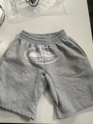 Grå sweatshorts med tryck - Säljer ett par grå sweatshorts med resår i midjan och ett vitt tryck på framsidan. Shortsen har avslappnad passform och är tillverkade i mjukt bomullsmaterial. Perfekta för chill dagar eller träning.