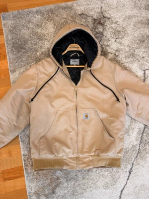  Carhartt OG active  - Snygg och efterbetraktad Carhartt OG active med dragkedja, svart quiltat innerfoder och två stora framfickor. Klassisk huva med svarta snören och ribbade muddar vid ärmslut och nederkant. Perfekt för dig som gillar streetwear och funktionell stil.
