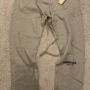 Grå Essentials mjukisbyxor - Säljer ett par grå mjukisbyxor från Essentials Fear of God. Byxorna har dragsko i midjan och muddar vid bensluten. Tillverkade i mjukt material för maximal komfort. Diskret logga på ena benet. Perfekta för en avslappnad stil.