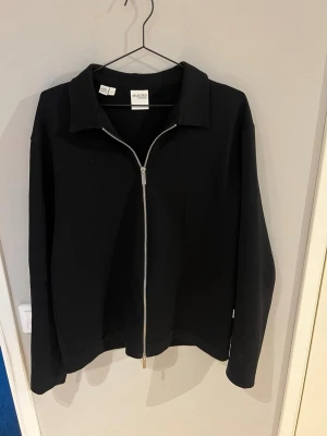 Svart Overshirt från Selected Homme - Stilren svart Overshirt från Selected Homme med klassisk krage och hel dragkedja framtill. Jackan har en enkel och clean design som passar perfekt till många olika outfits. Tillverkad i ett mjukt material som känns bekvämt hela dagen.