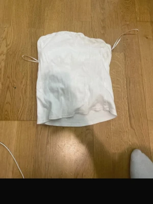 Vit ribbad bandeau topp H&M Divided - Säljer en vit bandeau topp från H&M Divided i ribbad bomull. Toppen är axelbandslös med tunna snören på sidorna och har en enkel, clean look. Perfekt till sommaren och lätt att matcha med olika outfits.