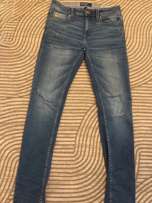 Blå slim fit jeans från Blend med slitningar  - Säljer ett par blå slim fit jeans från Blend med klassisk femficksdesign och snygga slitningar framtill. Jeansen har normal midja, smala ben och detaljerade bakfickor med broderi. Perfekta för en avslappnad och trendig look.