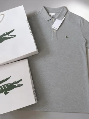 Grå pikétröja från Lacoste - Klassisk grå pikétröja från Lacoste med krage, knappar framtill och den ikoniska gröna krokodilloggan på bröstet. Tillverkad i mjuk bomull och har korta ärmar. Perfekt för en stilren och avslappnad look.