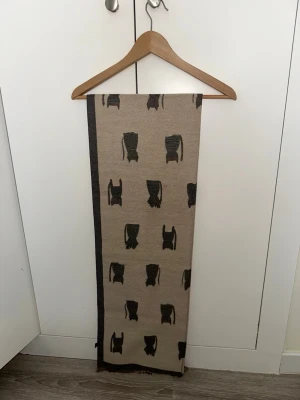 Beige halsduk med kattmönster - Säljer en beige halsduk med sött kattmönster i svart och grått. Halsduken är rektangulär och har en mörkare kant längs ena sidan. På andra sidan finns flerfärgade katter i orange, vitt och grönt. Perfekt för dig som gillar djurmotiv och vill ha en unik accessoar.