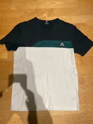 Le Coq Sportif färgblockad t-shirt - Snygg t-shirt från Le Coq Sportif med färgblockad design i svart, mörkgrönt och vitt. Klassisk passform med korta ärmar och rund hals. Ikonisk broderad logga på bröstet. Perfekt för en clean och sportig look.