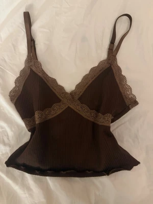 Vintage brun ribbad linnetopp med spets  - Brun ribbad linnetopp med tunna justerbara axelband och dekorativ spets längs urringningen och under bysten. Toppen har croppad passform och vågig kant nertill för en trendig look.