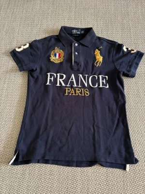 Mörkblå pikétröja Polo Ralph Lauren - Mörkblå pikétröja från Polo Ralph Lauren med broderad gul logga, FRANCE PARIS-tryck och detaljer i vitt och guld. Klassisk krage, kort ärm och knappar framtill. Perfekt för en sportig och stilren look.
