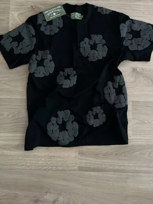 Denim tears t-shirt - Säljer en svart t-shirt från Denim Tears med stora gråa blommor tryckta över hela tröjan. T-shirten har rund hals och korta ärmar. Perfekt för dig som vill sticka ut med ett unikt mönster och cool design.