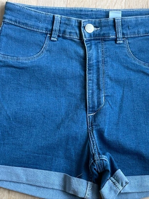 Blå denimshorts från H&M 164 - Klassiska blå denimshorts från H&M med upprullade benslut och femficksmodell. Shortsen har normal passform och bälteshällor i midjan. Perfekta för sommardagar och enkla att matcha med olika toppar.