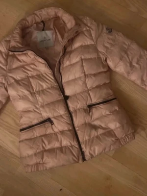 Moncler jacka - Snygg ljusrosa pufferjacka med svarta dragkedjor på fickorna och framtill. Jackan har quiltad design, hög krage och en badge på ärmen. Jag har använt den 1 gång!
