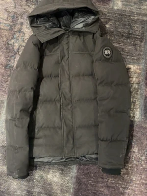 Canada goose macmillian - Snygg svart dunjacka med huva och quiltad design. Jackan har en patch på ärmen och dragkedja framtill med vindslå. Perfekt för kalla vinterdagar och har en stilren look som passar till det mesta.