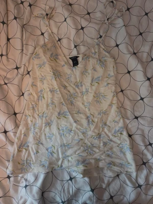 Blommig linnetopp från Lindex - Söt linnetopp från Lindex i ljusbeige med blått och grönt blommönster. Toppen har smala axelband med knyt och v-ringad hals. Perfekt för varma dagar och enkel att styla med jeans eller kjol.