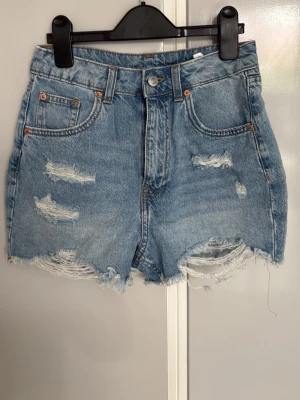 Högmidjade blå jeansshorts med slitningar - Säljer ett par högmidjade blå jeansshorts med slitna detaljer och fransig kant. Shortsen har klassisk femficksdesign, knappgylf och råa kanter nedtill för en avslappnad look. Perfekta för sommaren och ger en cool vibe till din outfit.