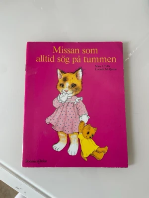 Missan som alltid sög på tummen - Barnbok med hårdpärm och starkt rosa omslag. Framsidan har en söt tecknad kattunge i rosa klänning som suger på tummen och håller i en gul nalle. Boken är ca 21 cm hög och utgiven av Bonnier Carlsen.