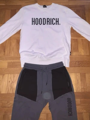 Hoodrich loungewear set i vitt och grått - Säljer ett loungewear set från Hoodrich med en vit sweatshirt med svart logga på bröstet och matchande gråa byxor med svarta detaljer och Hoodrich-tryck på benet. Perfekt för chill dagar hemma eller när du vill vara bekväm men ändå se snygg ut. Storlek S/M  hel sätt byxor o tröja perfekt för sommaren 