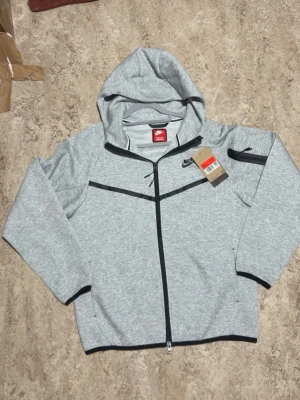 Ljusgrå Nike hoodie med dragkedja - Snygg ljusgrå hoodie från Nike med hel dragkedja framtill och svarta detaljer. Hoodien har en huva, svarta kantband vid ärmslut och nederkant samt en ficka med dragkedja på ena ärmen. Perfekt för en sportig och avslappnad stil.